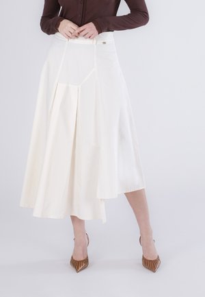 Kobieta ubrana w białą spódnicę midi z plisami oraz brązową bluzkę z długim rękawem, w zestawie z brązowymi szpilkami w szpic, stoi na tle jednolitego tła.