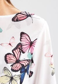 Haut en tissu fluide et blanc avec un imprimé de papillons en rose, bleu et noir. Le design comprend différentes tailles et formes de papillons.