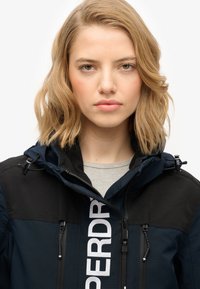 Granatowa kurtka z czarnymi akcentami, z zamkiem z przodu, kapturem i pionowym białym logo na piersi. Teksturowana tkanina z detalami w postaci sznurka.