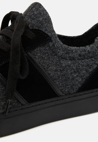 Šedá teniska s texturovanou tkaninou, černými semišovými akcenty a černými tkaničkami. Zaměření na detailní záběr konstrukce boty.