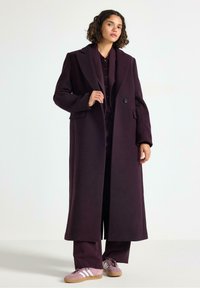 Lang, mørk lilla uldfrakke med krave, enkelt knap og sidelommer, båret over et matchende outfit og parret med pink sneakers.