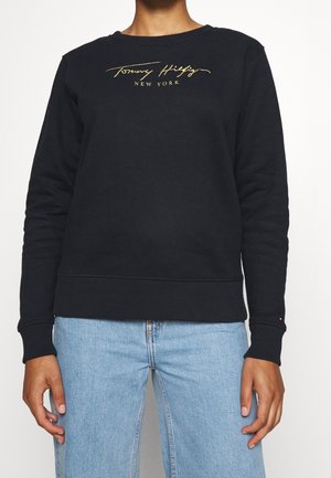 Personne portant un sweat-shirt noir à manches longues avec un texte doré "Tommy Hilfiger New York" et un jean bleu clair.