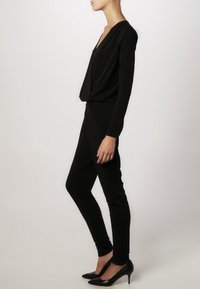Mono negro de manga larga, con escote en v drapeado, corte ajustado y piernas ceñidas. Combinado con tacones de aguja negros. Textura suave.