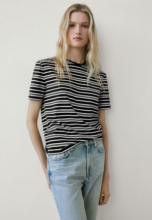 Femme blonde portant un t-shirt à manches courtes rayé noir et blanc et un jean bleu clair, debout contre un mur blanc uni.