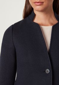 Marinblå texturerad kappa med trekantig krage, enkel knäppning och strukturerad form; bärs över en ljusbeige topp.