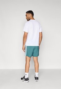 Feh�ér rövid ujjú atlétikai póló, türkiz rövidnadrággal, amelyen három fehér csík látható. A modell fekete sportcipőt és fehér zoknit visel.