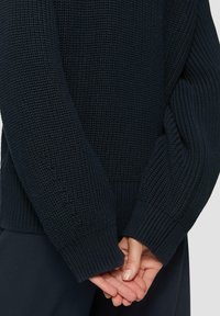 Maglione navy con lavorazione a trama, polsini a coste e vestibilità morbida, mostrato con le mani unite dietro la schiena.