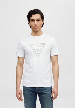 Un homme pose avec un T-shirt blanc orné du logo Guess et un jean bleu, sur un fond clair uni.