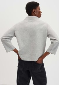 Grauer Pullover mit kraus gestricktem Kragen und 3/4-Ärmeln, kombiniert mit schwarzen, hochgeschnittenen Jeans. Weiche Textur, lässiges Design.