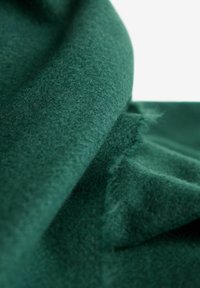 Temno zelena, mehka flis tkanina se zloži z vidno teksturo in rahlim ohlapljenjem na robovih pod svetlo osvetlitvijo.