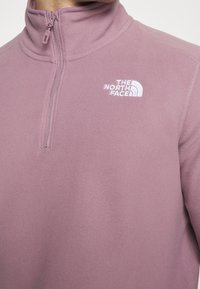 Lys lilla fleece pullover med halv lynlås, har en høj krave og broderet hvidt logo på venstre bryst. Blød tekstur.