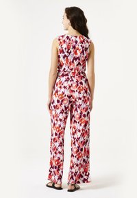 Mouwloze, bloemen jumpsuit met een riem in de taille, voorzien van een kleurrijk patroon in tinten van rood, paars en wit. Platte sandalen gedragen.