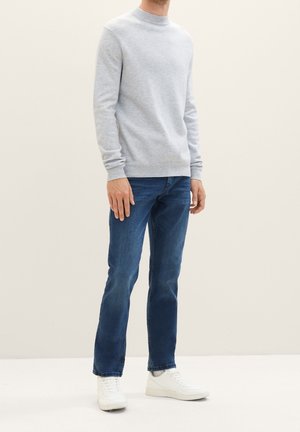 Hombre con suéter gris claro, jeans azules y zapatillas blancas de pie frente a un fondo neutro.