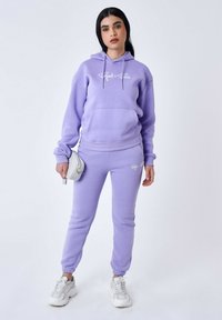 Paarse hoodie met trekkoorden en een voorzak, gecombineerd met bijpassende paarse joggers met een logo. Accessoires zijn een zilveren handtas.