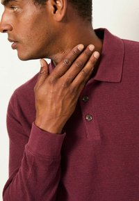 Burgunderfarbenes Poloshirt mit langen Ärmeln aus strukturiertem Stoff, mit einem Halsauschnitt mit drei Knöpfen und gerippten Bündchen, getragen von einer Person, die ihren Hals berührt.