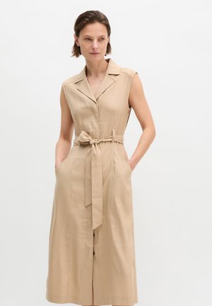 Frau in einem ärmellosen, beigen, mit Gürtel versehenen Kleid mit Kragen, Hände in den Taschen, schaut leicht zur Seite vor neutralem Hintergrund.