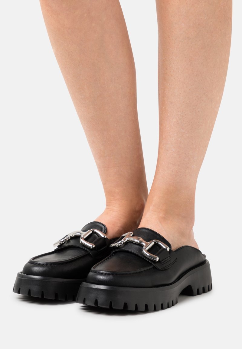 Sorte læder slip-on sko med en kraftig sål, der har sølvhardware detaljer og en rund tådesign. Tekstureret mønster under sålen.
