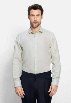 Mann mit kurzen dunklen Haaren und Bart, der ein hellgrünes langärmliges Hemd mit Knöpfen und dunkelblaue Hose trägt und vor weißem Hintergrund steht.