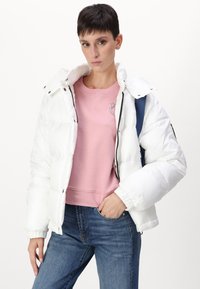 Witte gewatteerde jas met een glanzende textuur, voorzien van een hoge kraag en rits, gedragen over een roze sweatshirt en blauwe jeans.