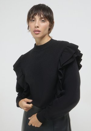 Maglione nero con maniche arricciate, scollo rotondo, texture morbida, maniche lunghe e polsini aderenti, indossato sopra una gonna nera aderente.