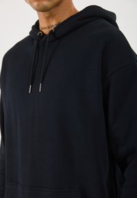 Svart hettegenser laget av myk stoff, med en frontlomme, snorhette og sølvfarget hardware-detaljer. Minimalistisk design uten mønstre.