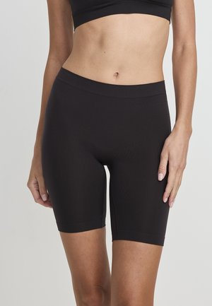 Frau trägt schwarze, hoch taillierte, enganliegende Biker-Shorts und ein schwarzes Crop-Top, steht vor einem schlichten hellgrauen Hintergrund.