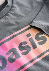Primer plano de una camiseta de tela gris con un parche rectangular con degradado rosa y naranja que muestra la palabra "Oasis" en letras negras en negrita.