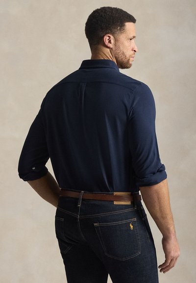 Muškarac u podvrnutoj mornarsko plavoj košulji i tamnim trapericama s smeđim remenom, prikazan straga na jednobojnoj bež pozadini.