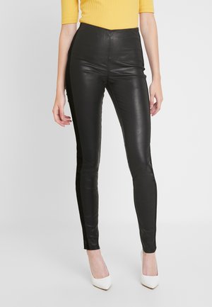 Hoge taille zwarte leren broeken met een strakke pasvorm, een gestructureerd oppervlak en contrasterende zijpanelen, gecombineerd met witte hoge hakken.