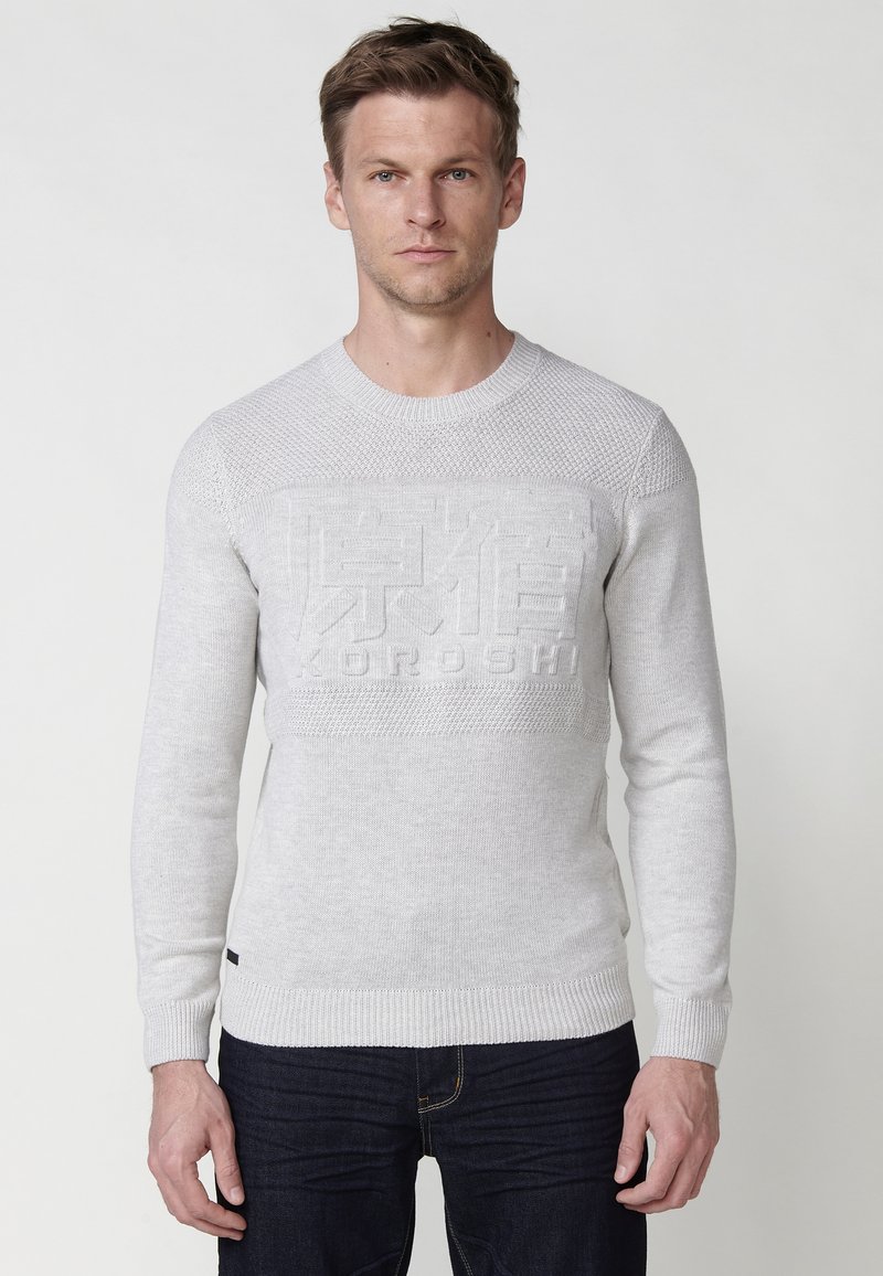 Lichtgrijze trui met een textuurpatroon, ronde halslijn en ribgebreide manchetten. Beschikt over een reliëfmerklogo op de voorkant. Jeans niet getoond.