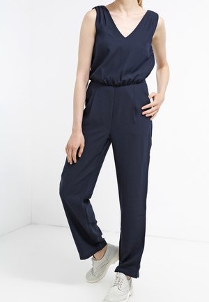 Mørkeblå jumpsuit med V-udskæring, ærmeløst design, elastisk talje og tapered ben, kombineret med lysfarvede sneakers.