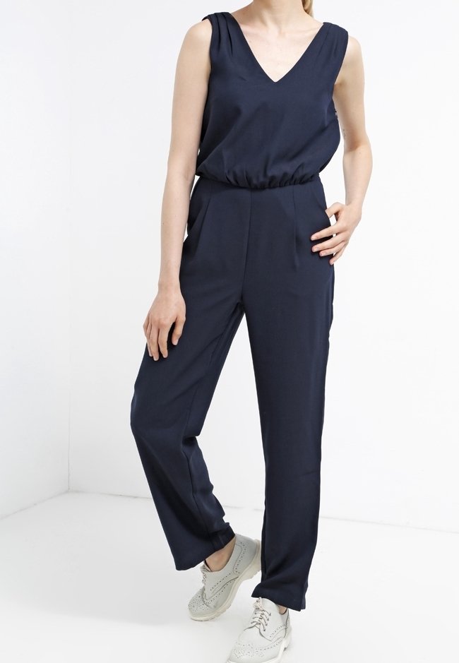 Jumpsuit bleu marine avec un décolleté en V, sans manches, taille élastique et jambes fuselées, associé à des baskets de couleur claire.