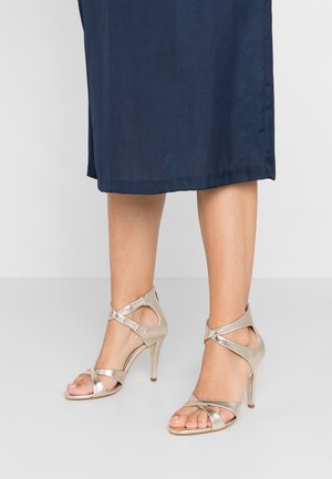 Złote sandały na wysokim obcasie z paskami, z krzyżującymi się paskami na kostce, otwartym noskiem i gładką fakturą. W połączeniu z ciemnoniebieską spódnicą.