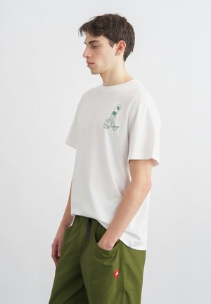 Giovane uomo con t-shirt bianca con piccolo disegno di una sneaker verde sul petto e pantaloni verde oliva, in piedi con le mani nelle tasche contro uno sfondo semplice.