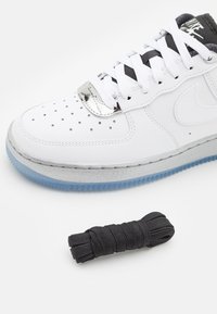 Baskets blanches Nike avec languette noire, lacets blancs et embout perforé, présentées avec une paire de lacets noirs séparée sur un fond blanc.