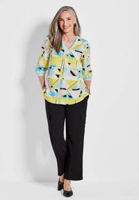 Bunte geometrische gemusterte Bluse mit Dreiviertelärmeln und V-Ausschnitt, kombiniert mit schwarzen weit geschnittenen Hosen und schwarzen Schuhen.