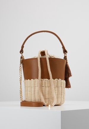 Brązowa skórzana torba typu bucket z plecionym rattanem, sznurkiem ze splotem warkocza, złotym łańcuszkiem jako paskiem oraz brązowym frędzlem na białym tle.