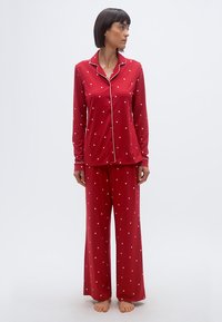 Femme debout pieds nus portant un pyjama rouge à manches longues avec des motifs de cœurs blancs, regardant vers sa gauche sur un fond uni.