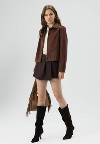 Bruine, textuurrijke jas met een kraag en knoopsluiting, gecombineerd met een donkere rok. Model draagt zwart, kniehoog, aansluitende laarzen en draagt een tas met franjes.