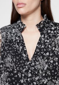 Schwarzblumiges Blusenshirt mit V-Ausschnitt und Rüschenausschnitt. Der Stoff ist leicht und hat ein strukturiertes, zerknittertes Muster mit weißen Blumenmotiven.
