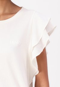 Balta T-krekls ar uzpūstām piedurknēm, ar smalku struktūras virsmu un mazu baltu logotipu "LIU.JO MILANO" priekšpusē.