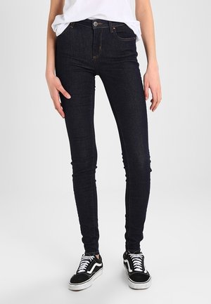 Vaqueros de tiro alto ajustados en denim azul oscuro, con un diseño clásico de cinco bolsillos y detalles de costura mínimos. Combinados con zapatillas negras.