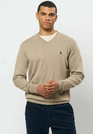 Mann trägt einen beigen V-Ausschnitt-Pullover über einem weißen Unterhemd und dunkelblauen Hosen, steht mit verschränkten Händen vorne.