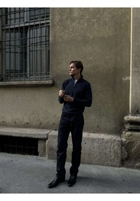 Mladý muž v čiernej košeli s dlhým rukávom a nohaviciach stojí na ulici a upravuje si rukávy, pričom stojí pred textúrovanou béžovou stenou s okenicami so zábranami.