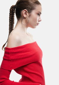 Czerwona, odkryta ramiona bluzka z prążkowanej tkaniny z długimi rękawami, prezentująca dopasowany krój i teksturowany materiał. Modelka nosi dużą srebrną kolczyk w kształcie koła.