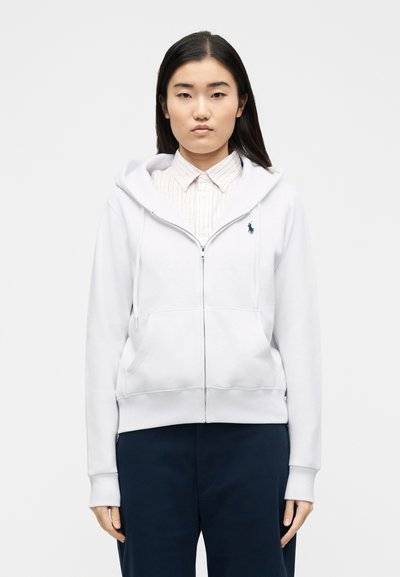 Giovane donna con indosso una felpa bianca con zip sopra una camicia a righe con colletto e pantaloni scuri, in piedi davanti a uno sfondo bianco uniforme.