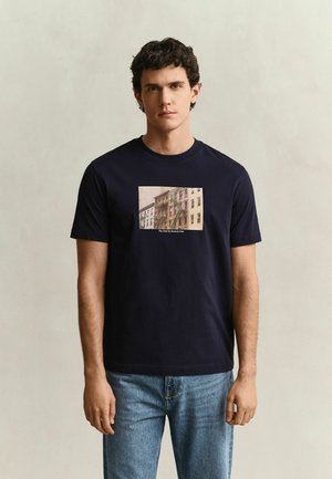 Jonge man met kort krullend haar draagt een marineblauw T-shirt met een stadsgebouw-grafiek en een blauwe spijkerbroek, staand tegen een effen lichte achtergrond.
