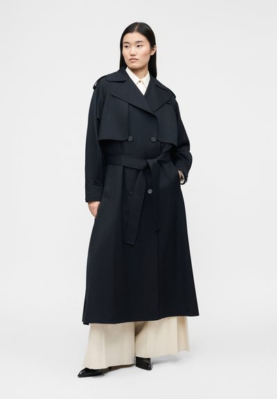 Femme debout portant un long trench-coat noir ceinturé sur un pantalon large crème et des chaussures noires pointues, sur un fond clair uni.