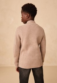 Pull en maille beige à texture côtelée, col montant et manches longues, assorti à un pantalon gris foncé. Fond aux tons neutres doux.