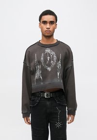 Μαύρο cropped φούτερ με λευκό γραφικό σχέδιο που παρουσιάζει περίπλοκα μοτίβα. Συνδυάζεται με σκούρο τζιν με φθορές και μαύρη ζώνη με καρφιά.